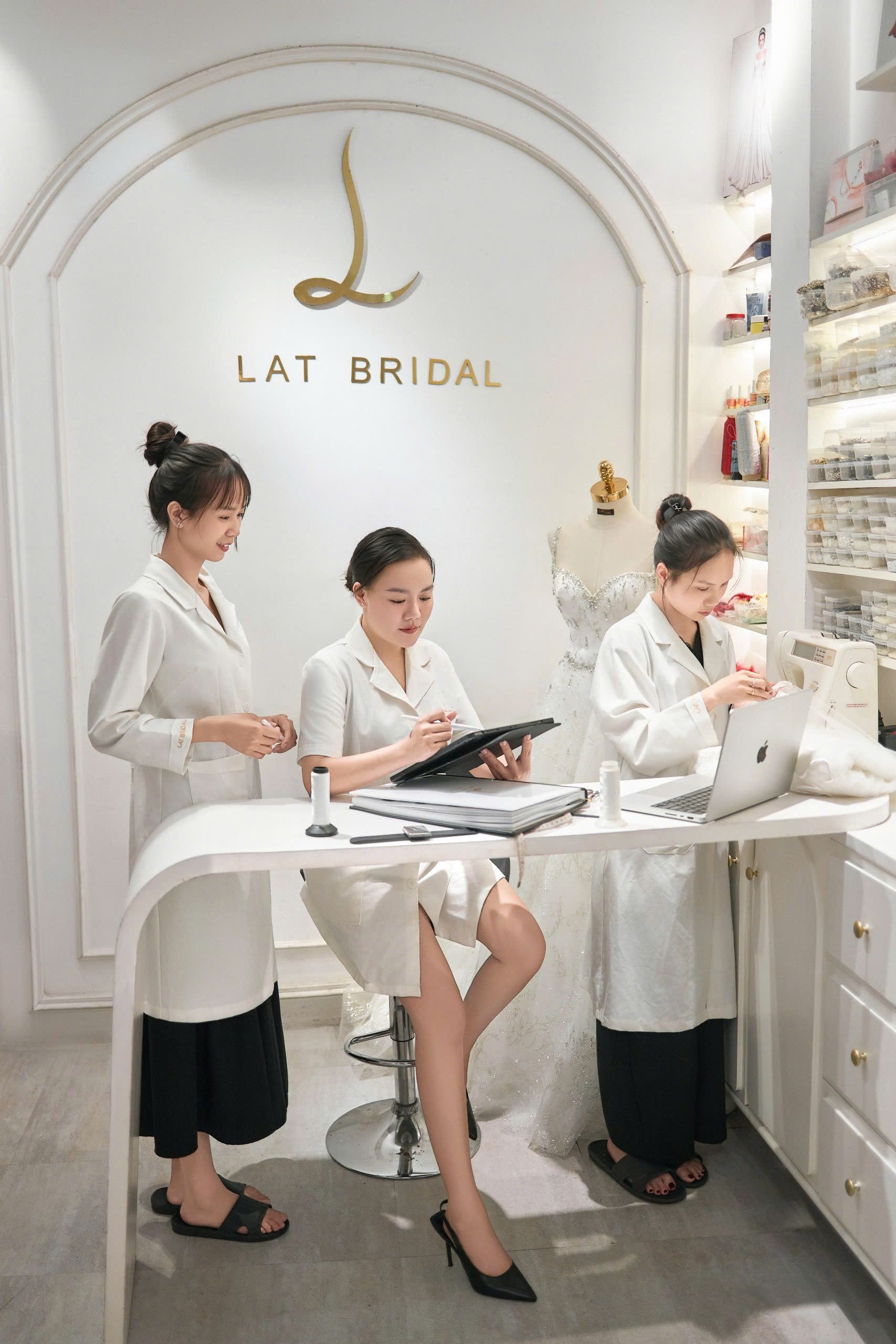 Ba người phụ nữ trong áo blouse trắng tại bàn của atelier, dưới bảng hiệu LAT Bridal